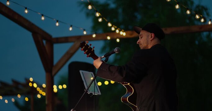 Male acoustic guitarist performing on outdoor evening stage under warm string lights