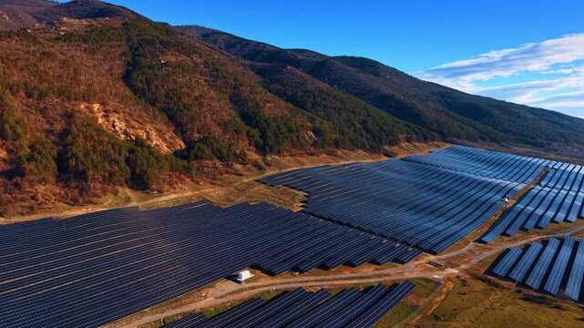 Solar farm nestled between wooded mountain ridges. Tiered panel sections on brown terrain with white inverter station and clear blue sky.
