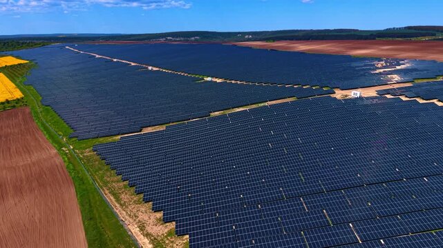 Massive solar installation surrounded by farmland and rapeseed fields. Flat terrain with dark panel rows, plowed brown soil and yellow crop patches under summer sky.
