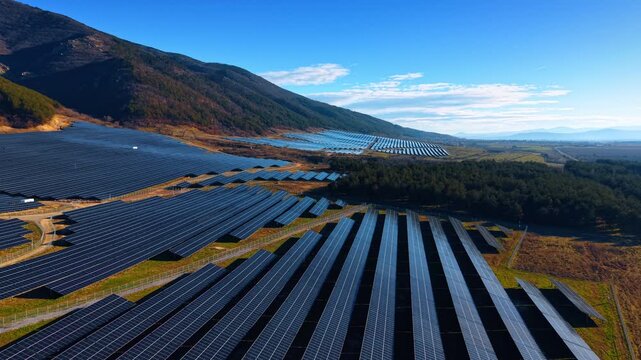 Solar arrays spanning mountain foothills. Rows extend from forested slopes into open plain with pine trees and scattered clouds.
