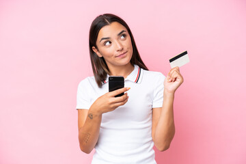Fototapeta premium Young caucasian woman isolated on pink background buying with the mobile with a credit card while thinking