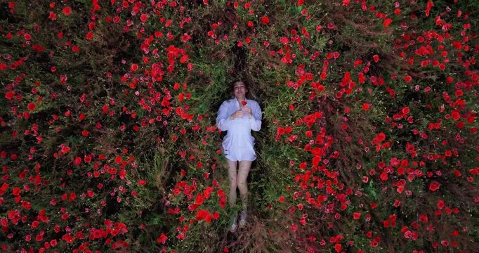 Young Woman Lying in Blooming Red Poppy Field Singing While Holding a Flower in White Dress Aerial Top View