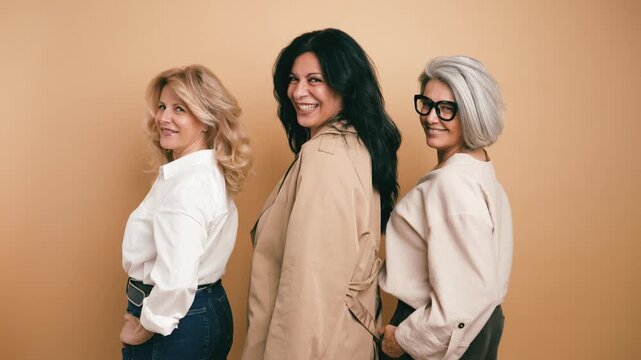 Three mature women of different generations with blonde, brunette, and gray hair turning to smile at the camera against a beige background