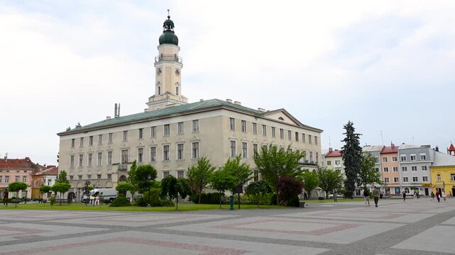 Main facade of the historic city hall building with a clock tower on the central Rynok Square in the old town of Drohobych, Western Ukraine, with pedestrians walking by on a cloudy summer day.