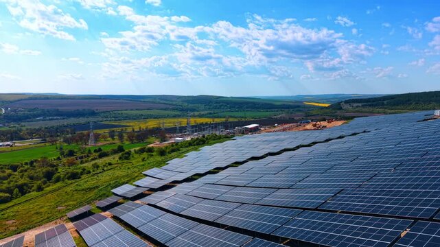 Solar park and electrical substation amid rolling green farmland. Panels stretch toward horizon with transmission towers, rapeseed patches and summer clouds.