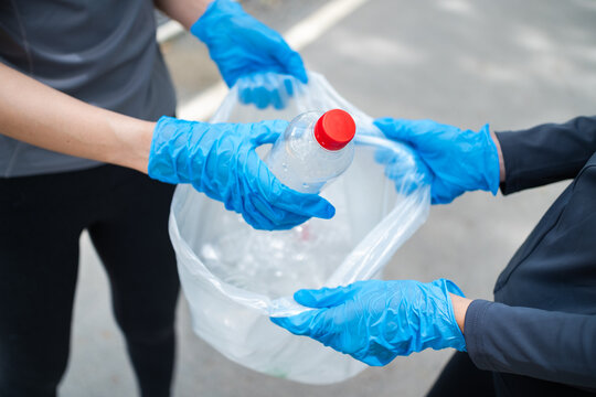 Two Asian women wearing blue gloves are collecting plastic waste. One person drops a water bottle into a trash bag to promote recycling, community service, and a cleaner environment.