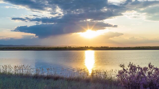 Clouds run across the sky with a sunset light over a lake with herbs on a shore
