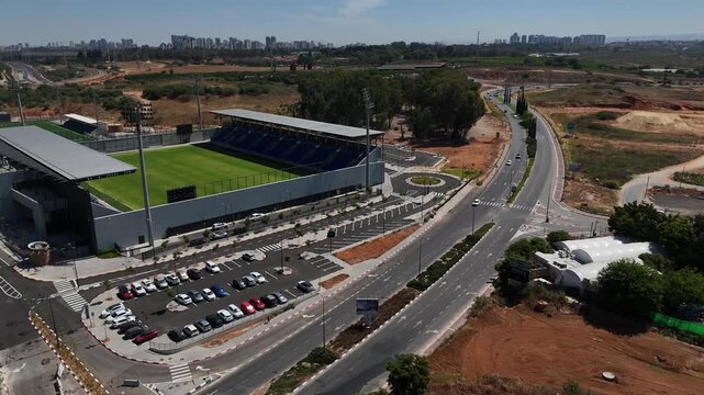 4K slow motion drone flight approaching the new stadium in Rehovot, revealing parking areas, eucalyptus grove and distant Ramla skyline, cinematic aerial view of urban development in Israel
