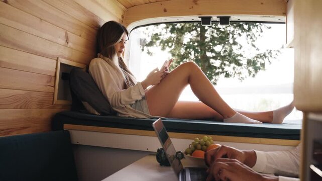 Young couple working as digital nomads inside their cozy camper van. The man is typing on a laptop, while the woman relaxes on the bed using her smartphone