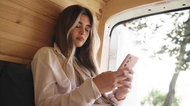 Beautiful young woman texting on her smartphone while sitting inside a camper van. She enjoys her road trip vacation, staying connected