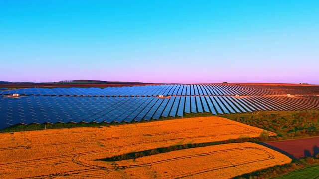 Large solar installation beside rapeseed and wheat fields at sunset. Orange horizon stretches behind colorful agricultural patchwork and blue panel arrays.