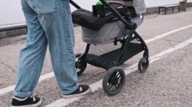 Close-up of a woman in jeans walking and pushing a baby stroller near the sea coast. The camera follows the legs and wheels to highlight the seaside promenade texture.