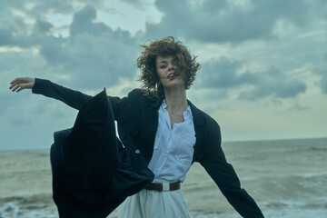 Fototapeta premium Windy beach fashion moment featuring a woman in a blazer and white shirt, coat sweeping in the breeze, sea horizon behind for a bold outdoor lifestyle and dynamic movement and energy