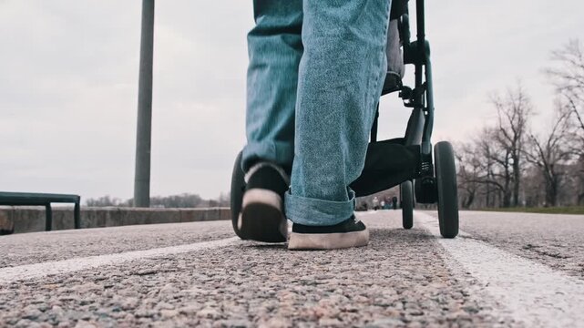 A woman pushes a stroller along a path bordered by grass and trees. The ground-level view captures the leisurely pace of the walk and the wheels moving on the pavement.