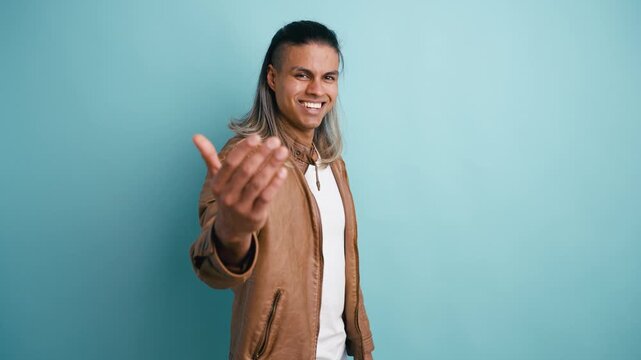 Happy and friendly young latino man in a brown leather jacket beckoning with a come here gesture and inviting to join on a blue background