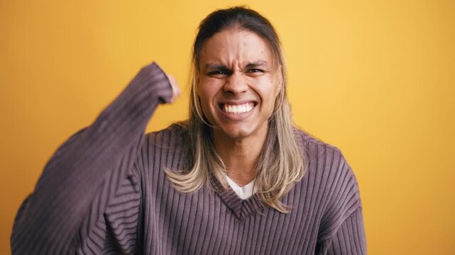 Happy long haired latino man celebrating victory with a joyful expression. He's clenching his fists in excitement in front of a yellow wall