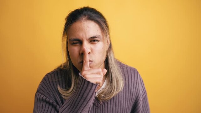 Latino man with long hair looking at the camera and putting a finger on his lips, asking to be quiet. Shhh gesture on a yellow background