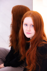 Naklejka premium Vertical portrait of pretty young blue-eyed girl with long wavy red hair and wearing black top and beige pants sitting next to her reflection in mirror 