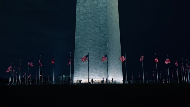 People walk around the Washington Monument in Washington, D.C. 555-foot-tall stone obelisk encircled by 50 American flags illuminated at night.