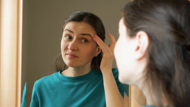Woman looking in mirror and examining wrinkles on her forehead and around eyes.