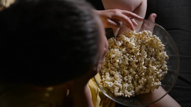 Top view of a young woman actively eating popcorn from a bowl. Focus stays on the bowl while her full body is visible in the frame, creating a casual snacking scene at home.