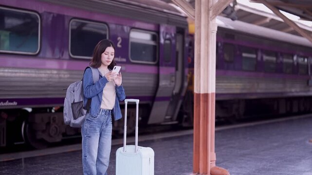 Young Asian woman traveler waiting on train platform with white suitcase and backpack checking smartphone before journey