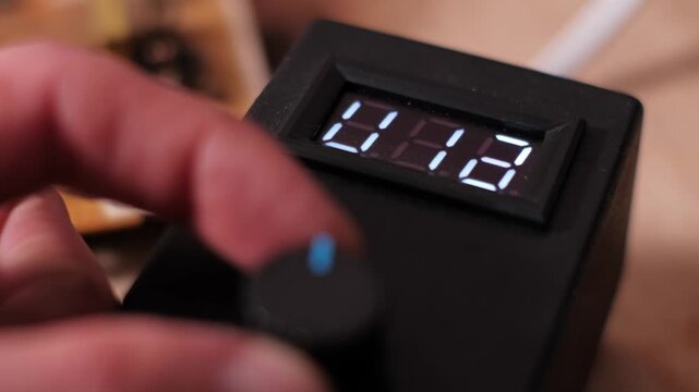 Close-up of a hand turning a rotary knob to adjust settings on an electronic control unit. The rapidly changing numbers emphasize precise calibration and power supply management.