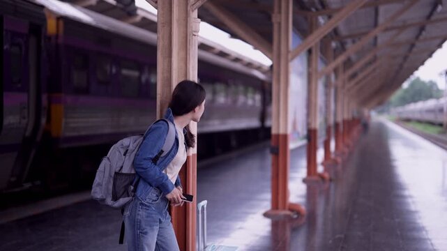 Young Asian woman traveler with backpack checking smartphone while waiting for train on outdoor railway platform during journey