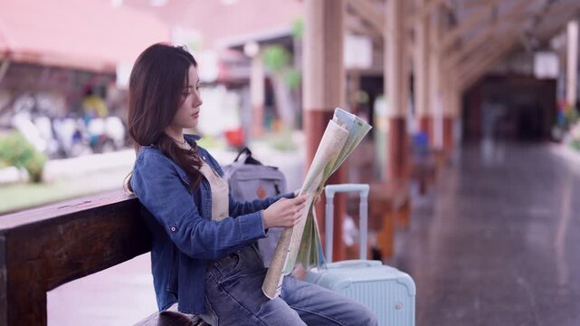 Young Asian woman traveler reading paper map while sitting with white suitcase at vintage train station platform waiting for departure