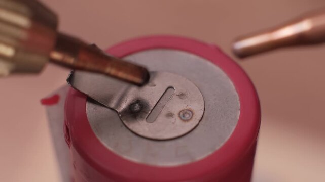 Extreme close-up of spot welder electrodes joining a nickel plate to a pink rechargeable battery. The precise operation illustrates modern energy storage manufacturing and assembly.