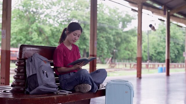 Young Asian female traveler reading book while sitting cross-legged on wooden bench at outdoor train station platform with backpack and blue luggage