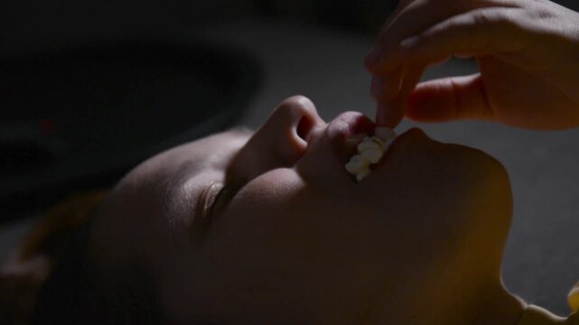 Top view close up of a young woman lying on her back and stuffing her mouth with large amounts of popcorn while watching a movie. 