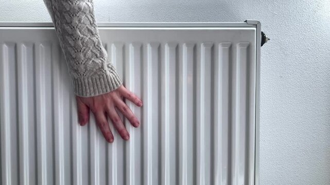 A woman checks the temperature of the radiator in the apartment, assessing the heat level and degree of heating. A woman's hand in a knitted sweater touches the heater