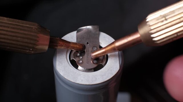 Close-up view of a spot welding process connecting a nickel strip to a grey lithium-ion battery. The copper electrodes against a dark background emphasize industrial precision.