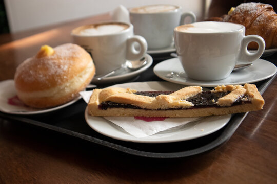 Vassoio con colazione italiana ricca composto da tre tazze di cappuccino con schiuma un bombolone alla crema un cornetto e una fetta di crostata alla marmellata su tavolo di legno