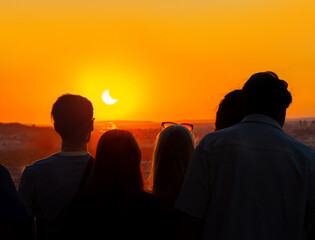 Silhouettes of people backlit by the light, observing a partial solar eclipse at sunset from the top of the Eiffel Tower. © sirbouman