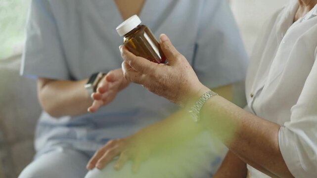 Professional caregiver or nurse explaining prescription medication instructions to an elderly senior woman holding a medicine bottle at home, providing supportive medical care in a high-quality 4K