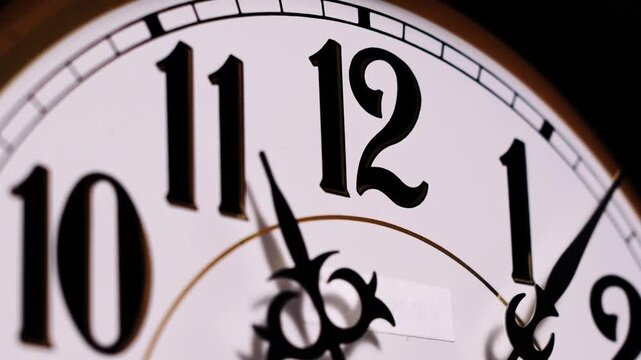 A close-up time lapse shows black clock hands spinning rapidly around a white dial with classic numerals towards twelve. Concept of looming deadlines, passing time, urgency, and the fast pace of life