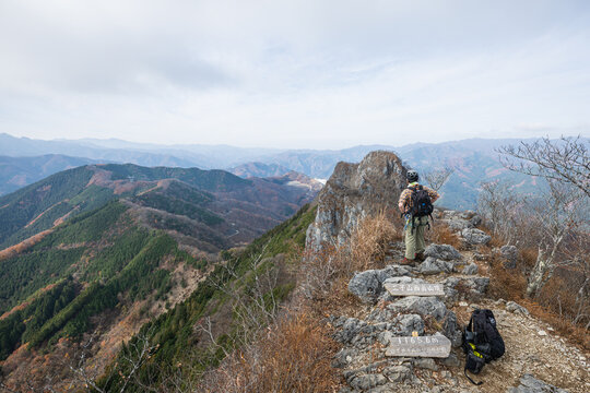Hiker standing on the rocky summit of Futago-yama West Peak at 1165.6m elevation with panoramic mountain landscape in Japan