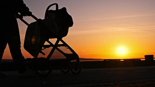 A dark silhouette of a parent pushes a baby stroller along a paved path against a vibrant orange sunset sky. Concept of peaceful parenting, evening family walks, and calm childhood moments.