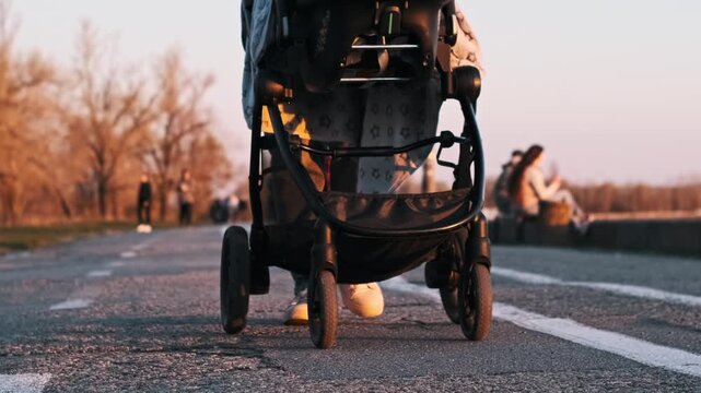 Close-up low angle shot of baby stroller wheels rolling on an asphalt path at sunset. The parent pushes the carriage through a park environment during the golden hour.