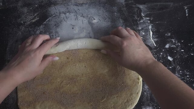 Top view of female hands rolling buttered dough covered with cinnamon and sugar filling into a tight log for homemade cinnamon rolls. Traditional baking process on a floured work surface