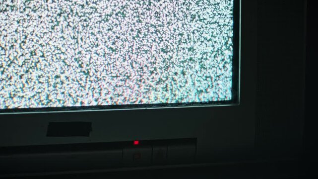 A close-up view of an old analog television screen flickering with static snow in a dimly lit room with a red light. Concept of signal loss, technical malfunction, and retro technology.