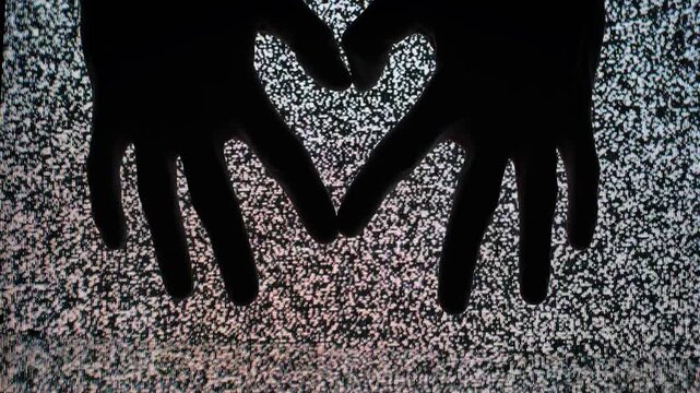 Silhouette hands form a heart shape gesture against a background of flickering television static noise, symbolizing love or connection amidst digital chaos or a message through the screen interference