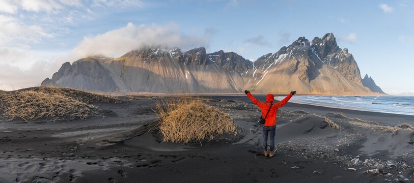 Man in red jacket stretches arms into the air, black lava beach, sandy beach, dunes with dry grass, mountains Klifatindur, Eystrahorn and Kambhorn, headland Stokksnes, mountain range Klifatindur, Austurland, East Iceland, Iceland