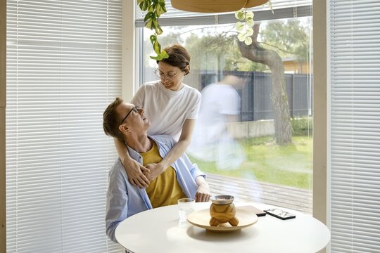 A couple shares a joyful interaction at a modern kitchen table. The woman embraces the man from behind while they engage in conversation, surrounded by natural light and green plants