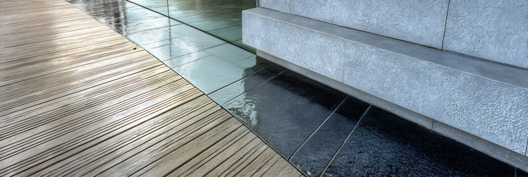 Modern architectural flooring transition combining grooved wooden decking and wet reflective dark stone tiles alongside a textured grey wall in a contemporary outdoor space
