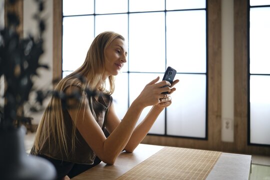 A woman sits at a wooden table in a room with large windows. She is focused on her phone, smiling softly. The room has natural light coming through the windows, creating a bright atmosphere