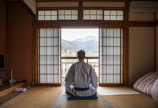 Young man wearing kimono sitting in traditional Japanese living room with tatami mats and shoji sliding doors, from behind, Yamanouchi, Nagano, Japan