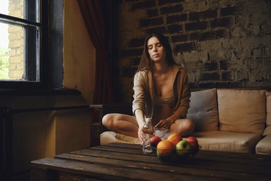 A pregnant woman is sitting on the couch near a window and reaching for a bottle of water on the table